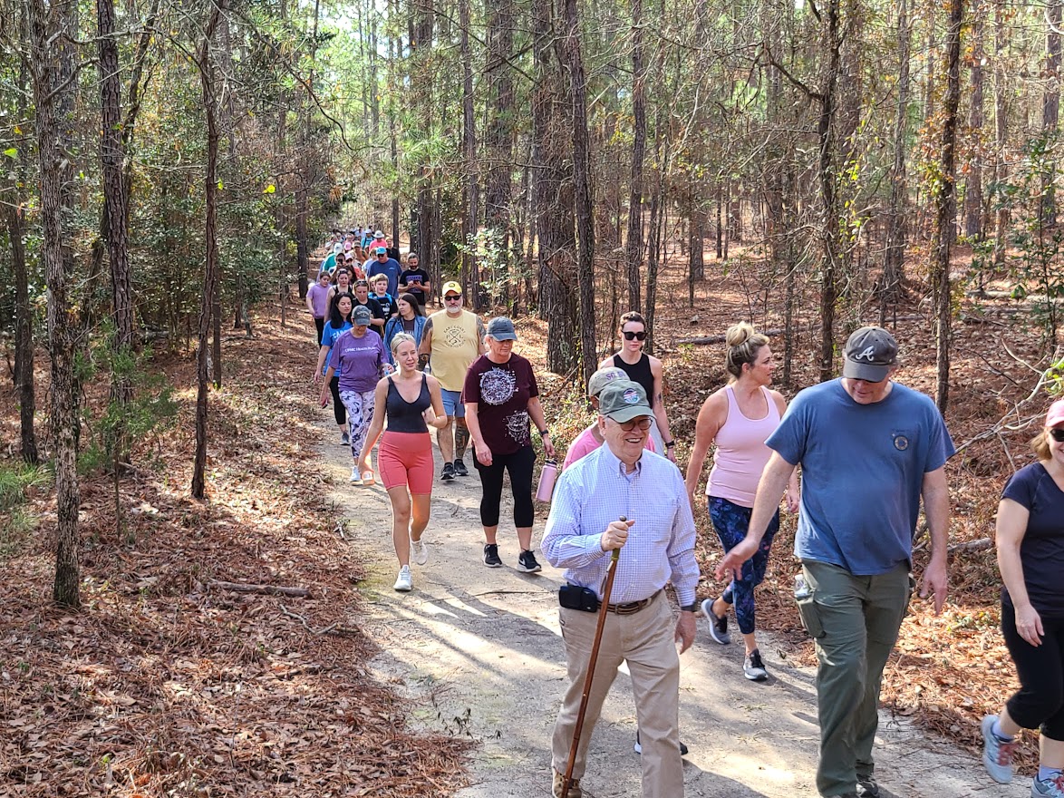 A parade of people walking the Goodale state park Nature trail on The First Day Hike. One person with a walking stick and people in comfortable clothing smiling