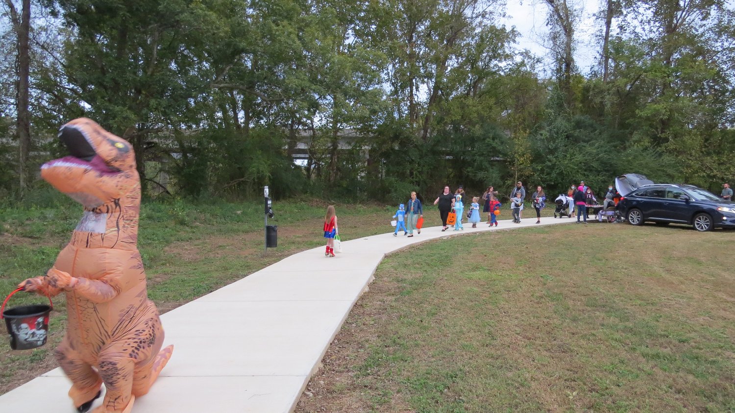 A parade of small children in Halloween Costumes getting out of cars going trick or treating. An adult sized dinosaur is in the foreground trick-or-treating at the Veterans Park Loop Trail