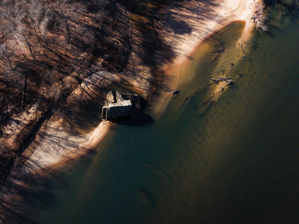 Aerial view of a shoreline with trees, clear water, and a large rock or concrete structure near the water’s edge.