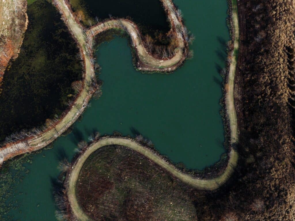 A winding dirt path separates two greenish ponds, surrounded by sparse trees and brown grass, viewed from above.