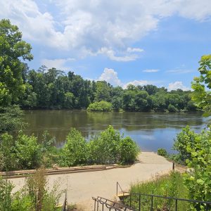 A concrete boat ramp leads to a calm, tree-lined river under a partly cloudy blue sky.