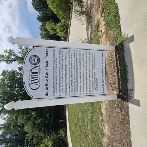 White sign with rules for using the Camden, SC boat ramp and water trails, surrounded by trees and pathway.