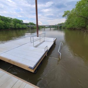 A floating dock with a handrail on a river, trees on both sides, and a bridge in the background.