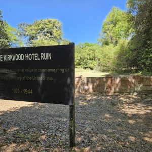 A sign reads The Kirkwood Hotel Ruin in front of old brick ruins surrounded by trees under a blue sky.