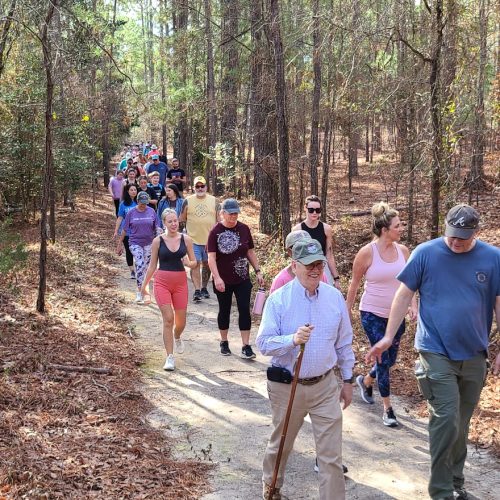 A parade of people walking the Goodale state park Nature trail on The First Day Hike. One person with a walking stick and people in comfortable clothing smiling