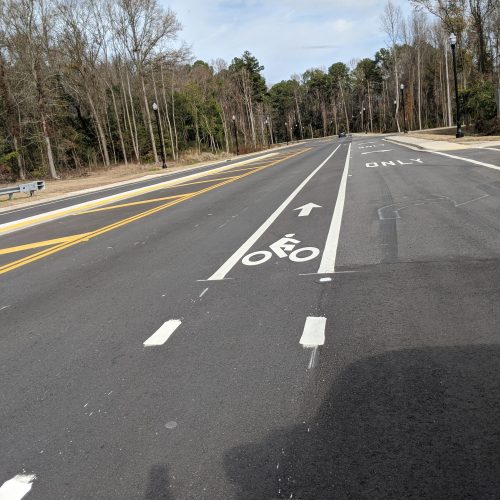 Empty street with a marked bike lane, lined by trees and streetlights under a partly cloudy sky.