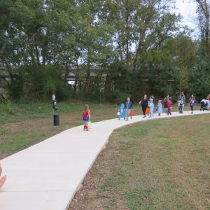 A parade of small children in Halloween Costumes getting out of cars going trick or treating. An adult sized dinosaur is in the foreground trick-or-treating at the Veterans Park Loop Trail